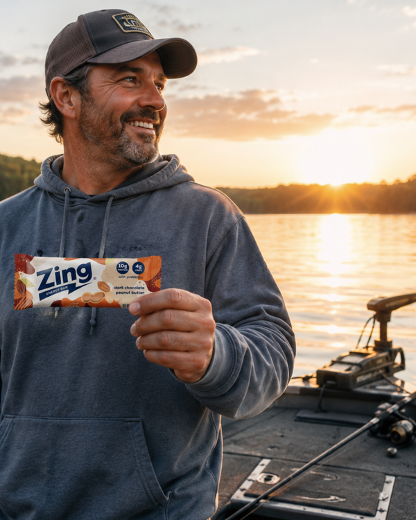 man holding a dark chocolate peanut butter zing energy bar while standing beside a lake at sunrise