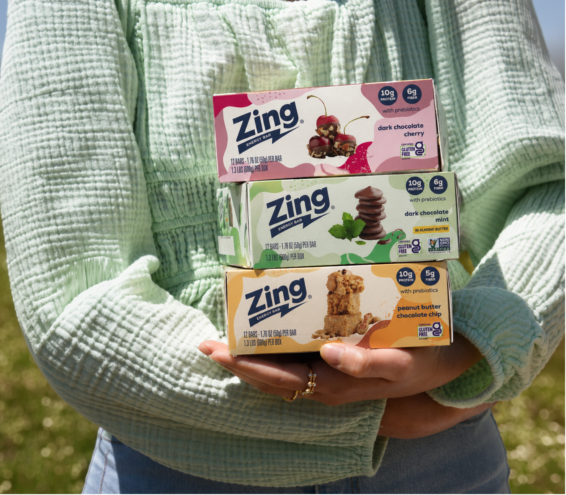 Person holding Zing Bars Dark Chocolate Cherry, Dark Chocolate Mint, and Peanut Butter Chocolate Chip boxes outdoors